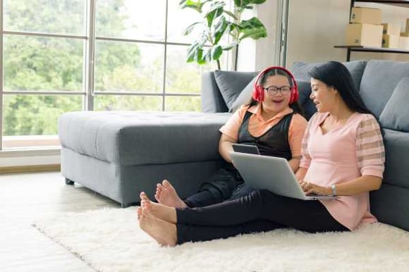 2 women sitting on a rug with their backs against a couch, looking at a laptop