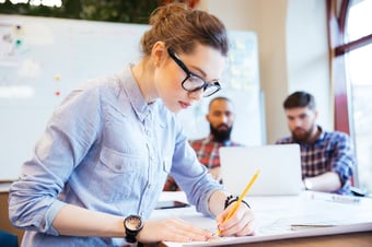 Focused engineer working on blueprints in an office setting with colleagues in the background