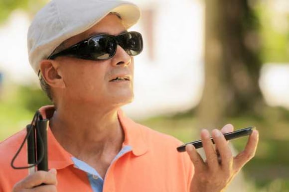 a man with a hat on, orange polo shirt, and holding a collapsable white cane in one hand holds a mobile phone in the other