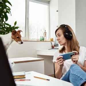 a woman at her desk, legs on it, headphones on, holding her smartphone, looking at a dog