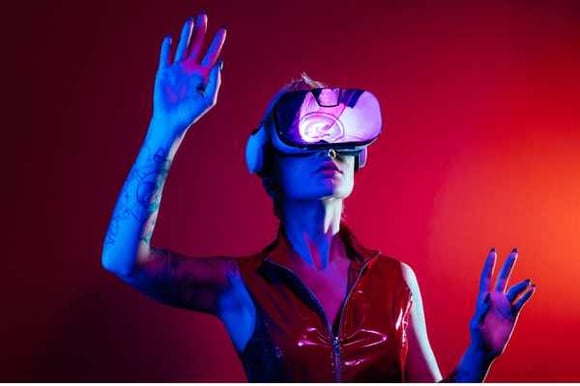 a woman in front of a bright red background with AI Goggles on, looking up, with her hands outstretched