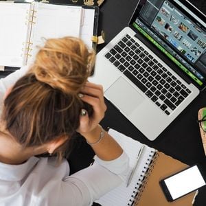 a woman in front of her laptop holding her head in her hands in apparent frustration with notebooks all around her