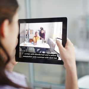 a woman watching a video on a tablet of two people with an audio description on the bottom of the video