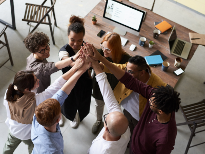 a-diverse-team-of-8-stand-in-a-circle-with-one-arm-from-each-outstretched-to-the-center-clapping-hands-together-in-front-of-a-desk-and-monitor