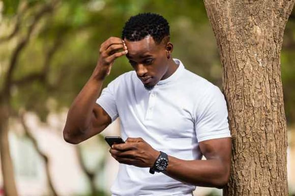a-man-next-to-a-tree-with-a-white-polo-on-looking-at-atext-message-on-his-phone-while-scratching-his-head