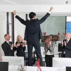 four_business_people_stand_around_clapping_while_one_business_man_with_his_back_to_the_camera_stands_on_a_desk_with_his_arms_in_the_air_in_celebration