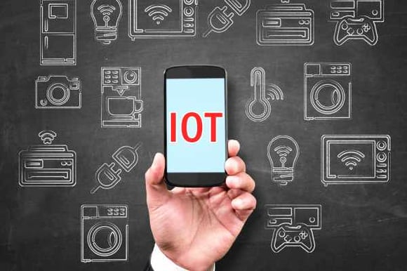 hand-holding-smartphone-with-IOT-displaying-in-text-in-front-of-a-blackboard-with-various-IOT icons