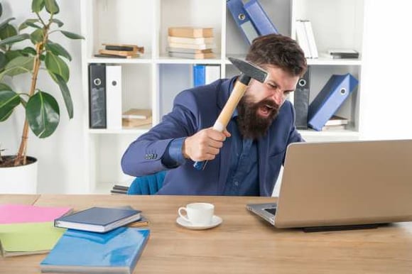 man-with-a-hammer-pointed-at-his-laptop-on-a-desk