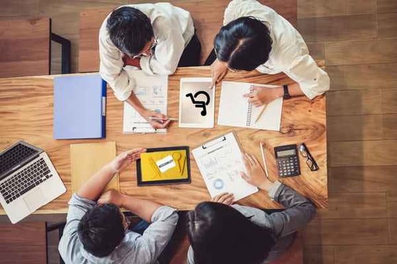top_down_view_of_four_people_at_a_wood_table_examining_tablets_and_laptops_with_report_printouts_scattered_on_the_table
