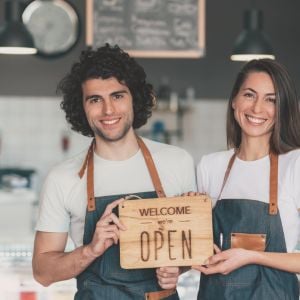 two coworkers in aprons jointly holding a sign that says welcome were open
