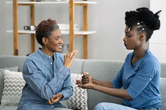 two women sitting on a couch, communicating with ASL, one is smiling and the other has a stern look on her face